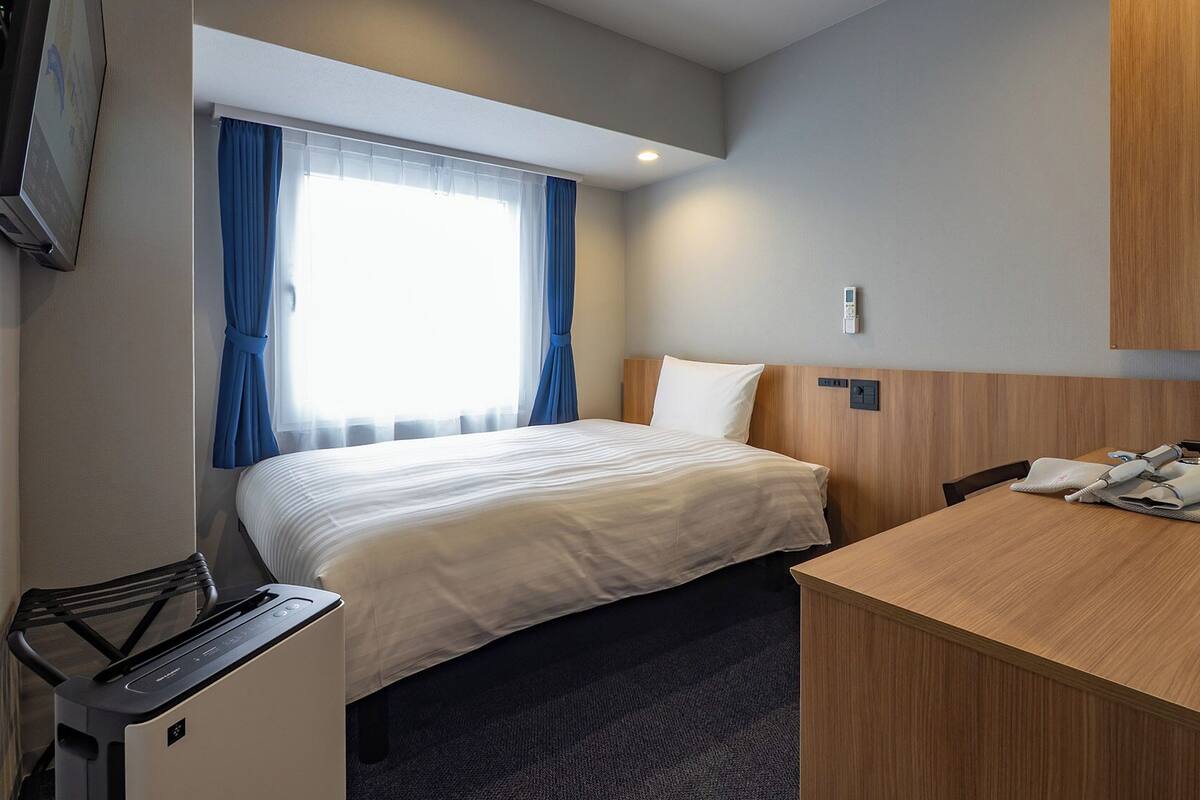 Quarto de hotel com cama, escrivaninha, cadeira, televisão e janela com cortinas.