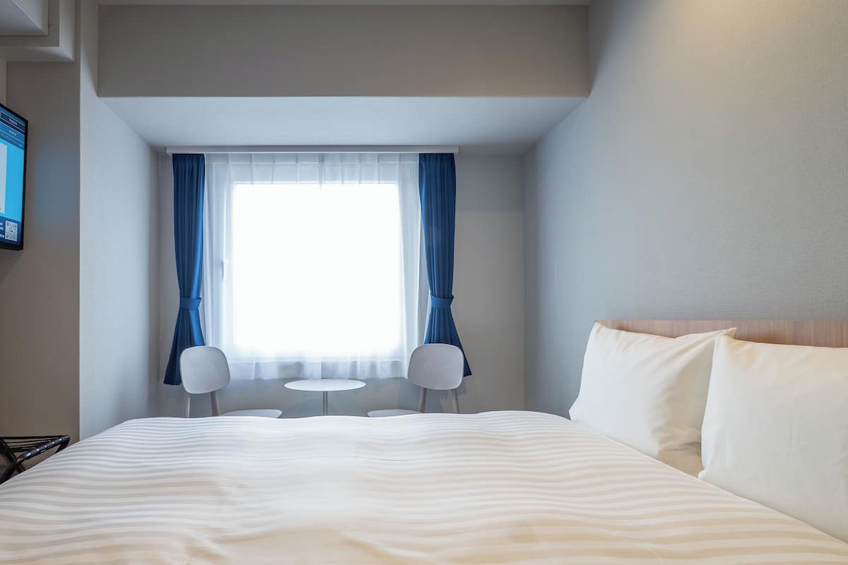 Quarto de hotel com uma cama grande, duas cadeiras, uma mesinha, televisão e uma janela com cortinas.