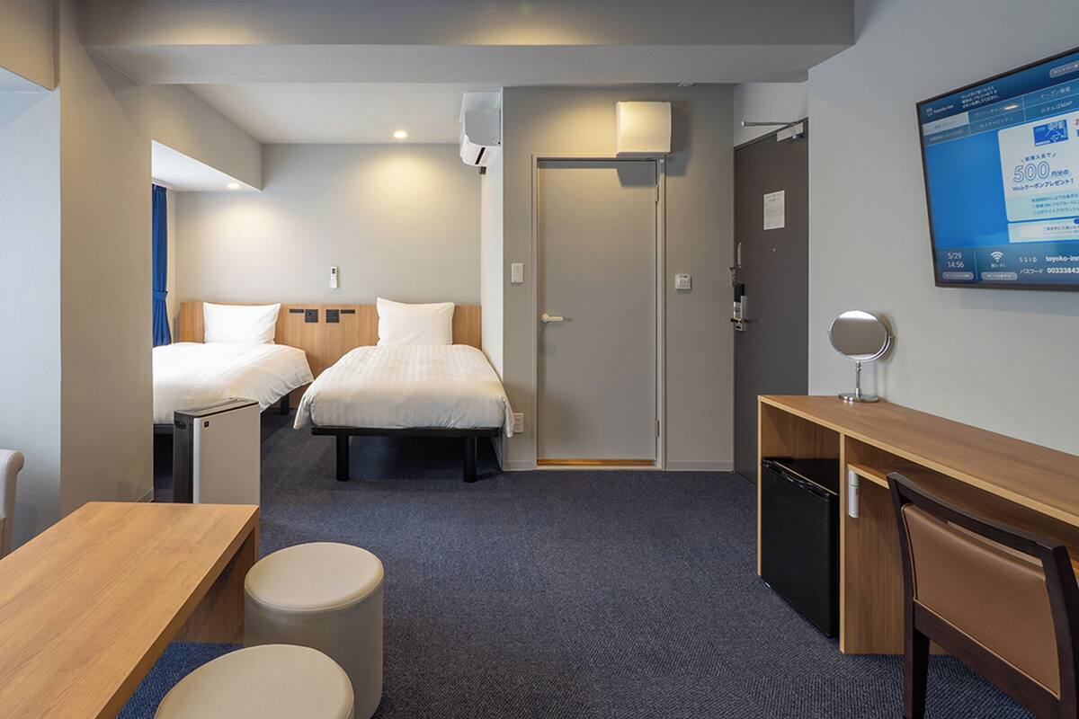 Quarto de hotel com duas camas, uma escrivaninha, uma cadeira, uma mesinha, um cesto de lixo e uma televisão na parede.