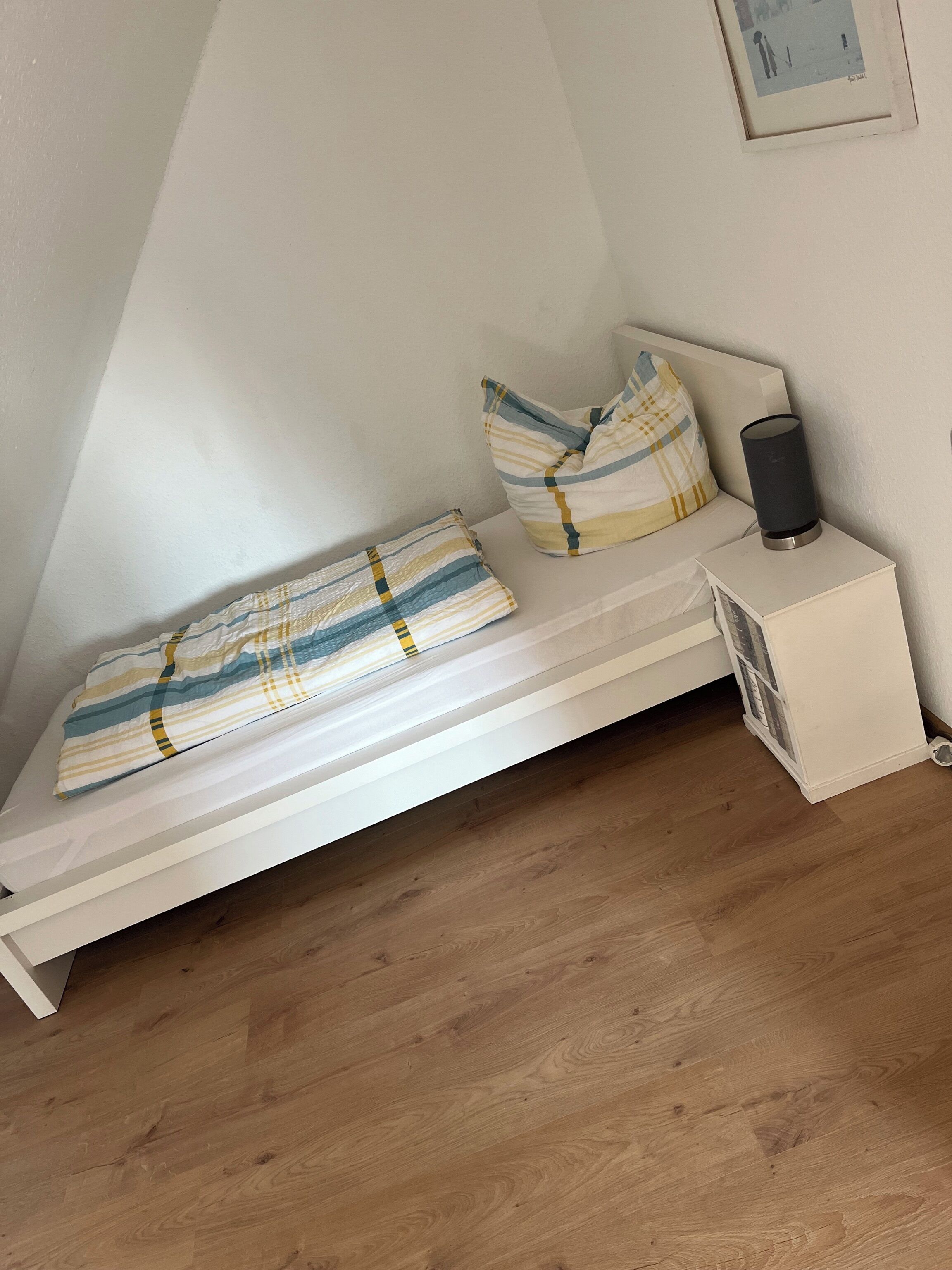 Ein kleines Zimmer mit einem Einzelbett, weißen Wänden, Holzboden, einem Nachttisch mit Lampe und einem gerahmten Bild an der Wand.