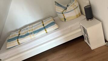Ein kleines Zimmer mit einem Einzelbett, weißen Wänden, Holzboden, einem Nachttisch mit Lampe und einem gerahmten Bild an der Wand.