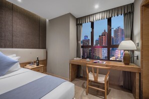 Una camera d'albergo con un letto, una scrivania con una sedia, un comodino, una lampada e vista sugli edifici della città.
