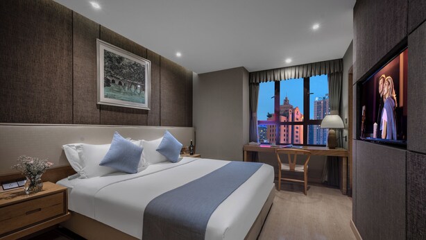 Ein Hotelzimmer mit einem großen Bett, einem Schreibtisch, einem Stuhl, einem Fernseher und einem Fenster mit Blick auf die Stadt.
