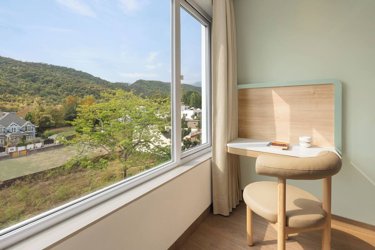 A room with a view of a green landscape and houses, a small wooden desk, and a chair.