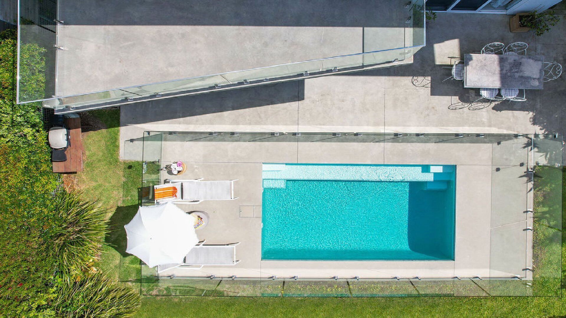 An aerial view of a swimming pool with a glass enclosure, surrounded by a patio area with outdoor furniture and a small garden.