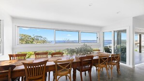A dining area with a large table and chairs, large windows with a view of the sea and greenery, and a glass door leading to an outdoor area. - Stunning ocean views and pool (KILLCARE HEIGHTS)