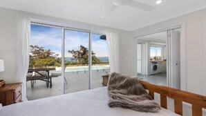 A bedroom with a large bed, a view of a pool and outdoor area, and a kitchen visible through an open door. - Stunning ocean views and pool (KILLCARE HEIGHTS)