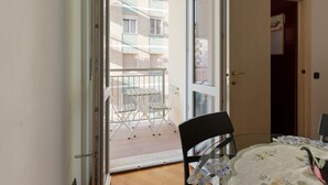 Une porte ouverte donnant sur un balcon avec une table et des chaises, d’où l’on peut admirer un bâtiment, et une autre porte menant à une autre pièce.