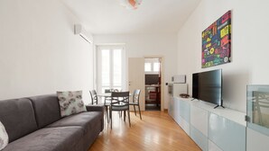 Un salon moderne avec un canapé gris, une table à manger avec des chaises, un téléviseur posé sur un meuble et une œuvre d’art colorée accrochée au mur.