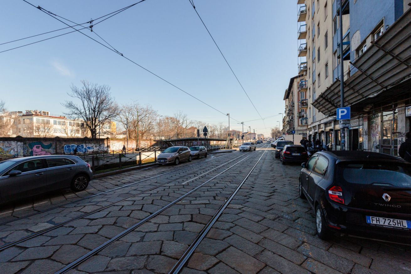 Straßeneisenbahn-Gleis mit parkenden Autos, einem Parkschild und Häusern mit Balkonen.