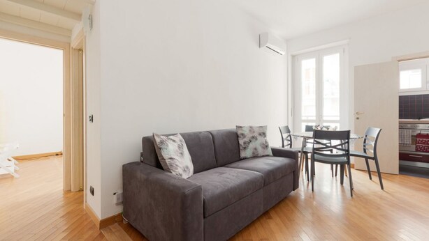 Un salon moderne avec un canapé gris, une table à manger avec des chaises et un coin cuisine visible à travers une porte ouverte.