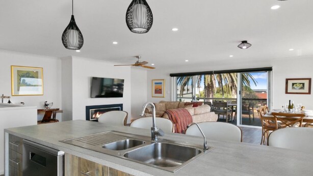 A modern kitchen with a central island, built-in oven, and stainless steel sink. There is a dining area with a table and chairs, and a living space with a sofa and a fireplace.
