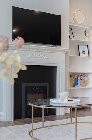 Cozy living room with a flat-screen TV above a fireplace, a glass table, and a bookshelf.