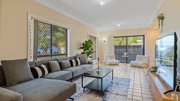 A living room with a sofa, coffee table, and chairs. There are large windows and a sliding door leading to a balcony.