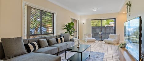 A living room with a sofa, coffee table, and chairs. There are large windows and a sliding door leading to a balcony.
