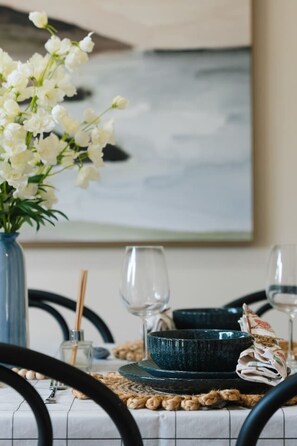 Une table à manger dressée avec un saladier bleu, des verres à vin et un vase contenant des fleurs blanches.