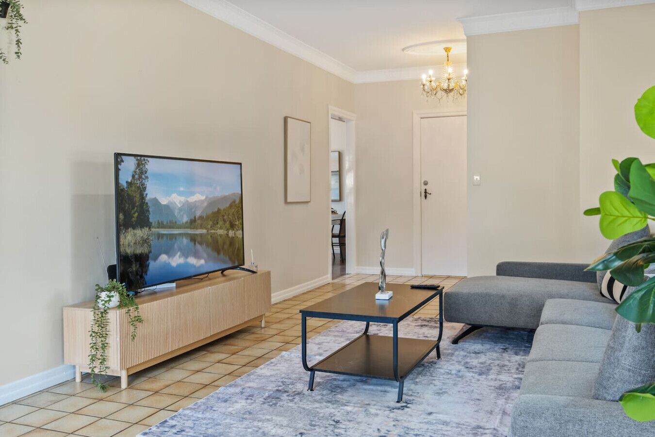 A living room with a flat-screen TV, a wooden cabinet, a coffee table, a grey sofa, and a large green plant.