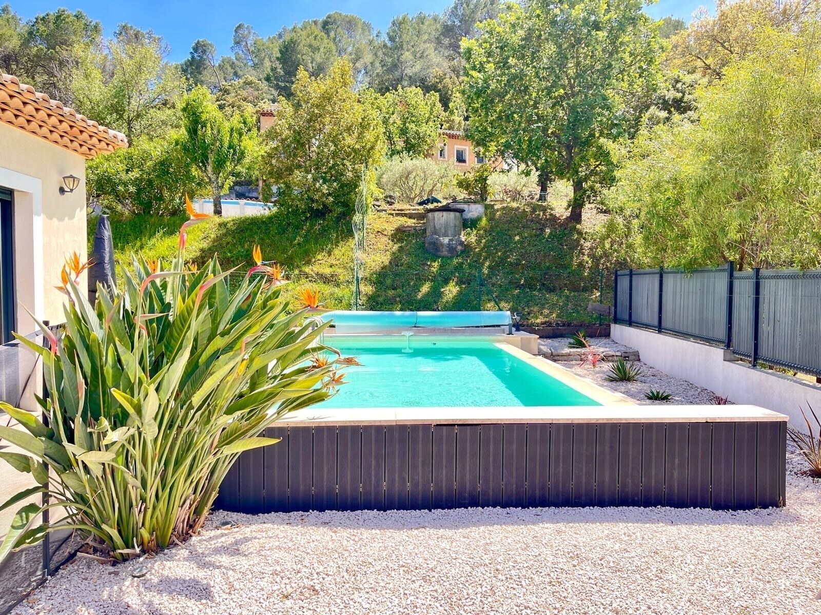 Une piscine avec une terrasse en bois, entourée de gravier et de plantes, avec des maisons et des arbres en arrière-plan.