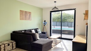 Ein modernes Wohnzimmer mit einem grauen Ecksofa, einem kleinen Couchtisch und einem Koffer. Ein Glastür öffnet sich zu einem Außenbereich mit einem Pool.