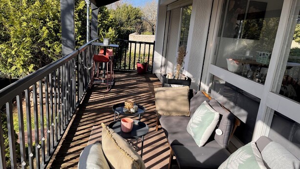 A sunlit outdoor seating area with a wooden deck, cushioned seating, and a view of trees and a stone wall. - Spacious country house, 15 min from Bordeaux city center (Latresne)