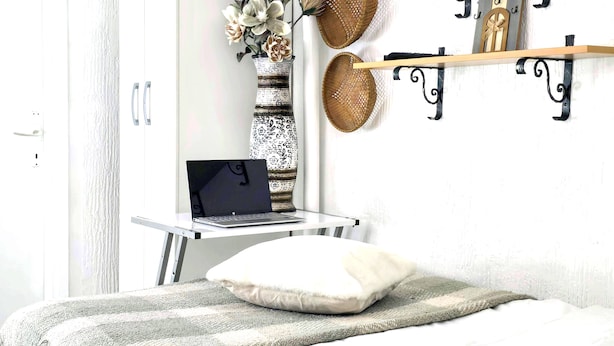 A bedroom with a bed, a laptop on a small table, a decorative vase with flowers, and wall-mounted shelves with baskets and figurines. - Room-angelina Apartment 3 (Belgrade)