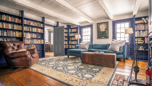 Een gezellige woonkamer met houten vloeren, een blauwe bank, een leren fauteuil en een grote boekenkast vol boeken.