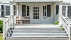 Een wit huis met een veranda, een houten bank en een trap die naar de ingang leidt.