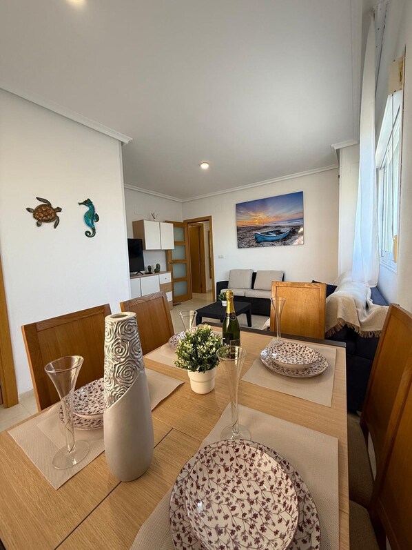 A modern living room with a dining area, featuring a sofa, a coffee table, and a painting of a sunset.