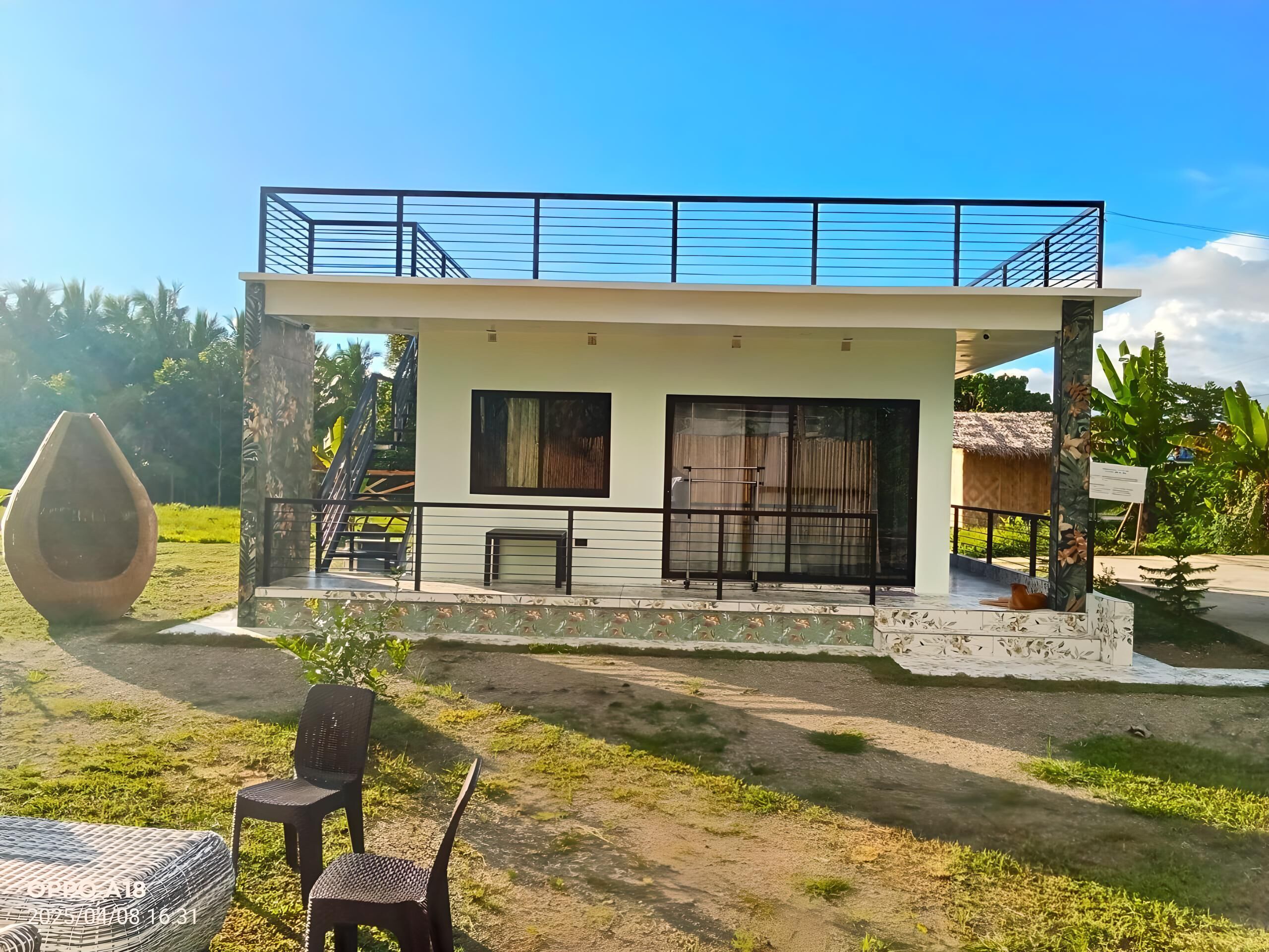 A modern two-story house with a balcony, a large window, and a patio area with chairs.