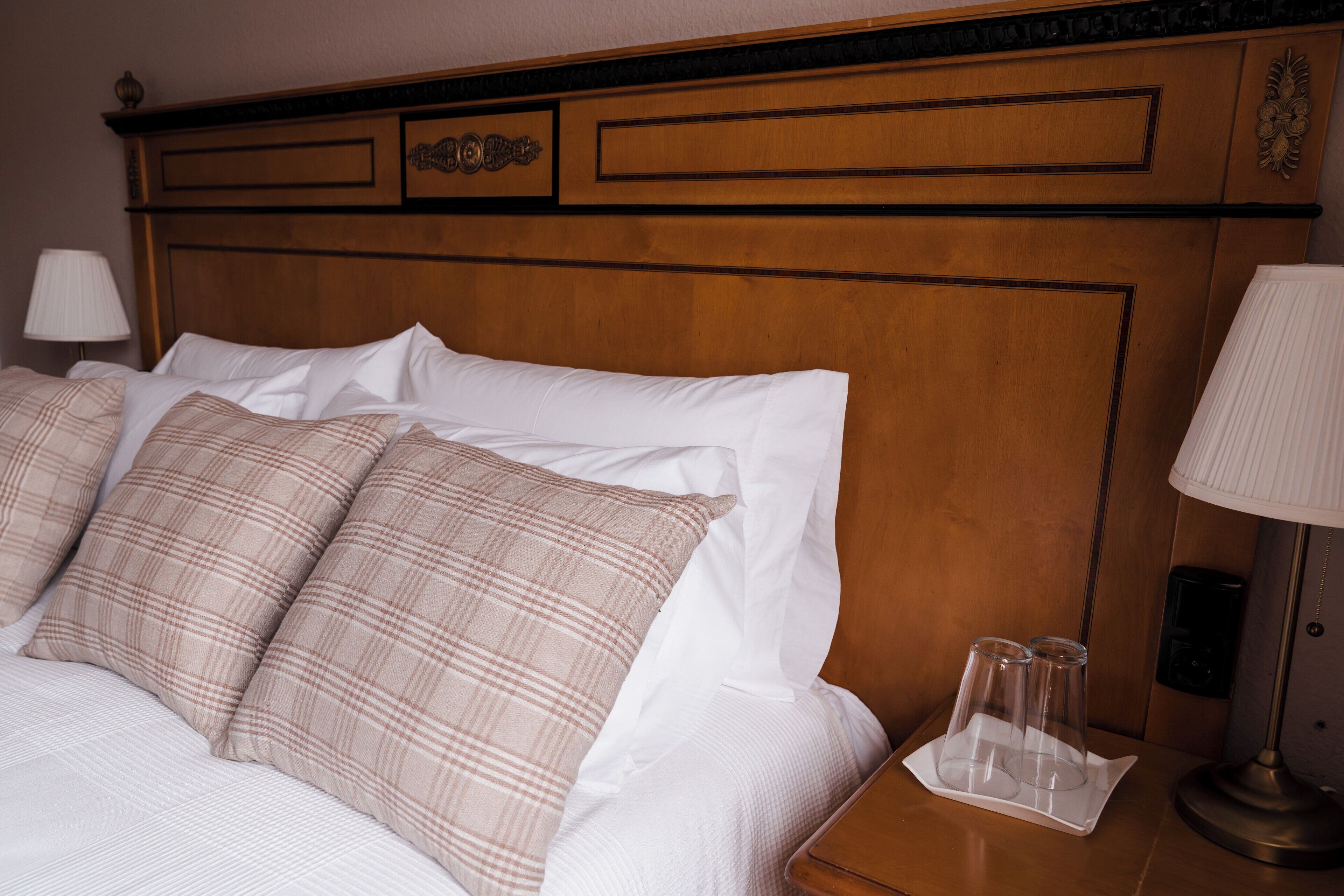 Una cama bien hecha con almohadas de cuadros y una mesita de noche con una lámpara y un reposabajares con vasos.