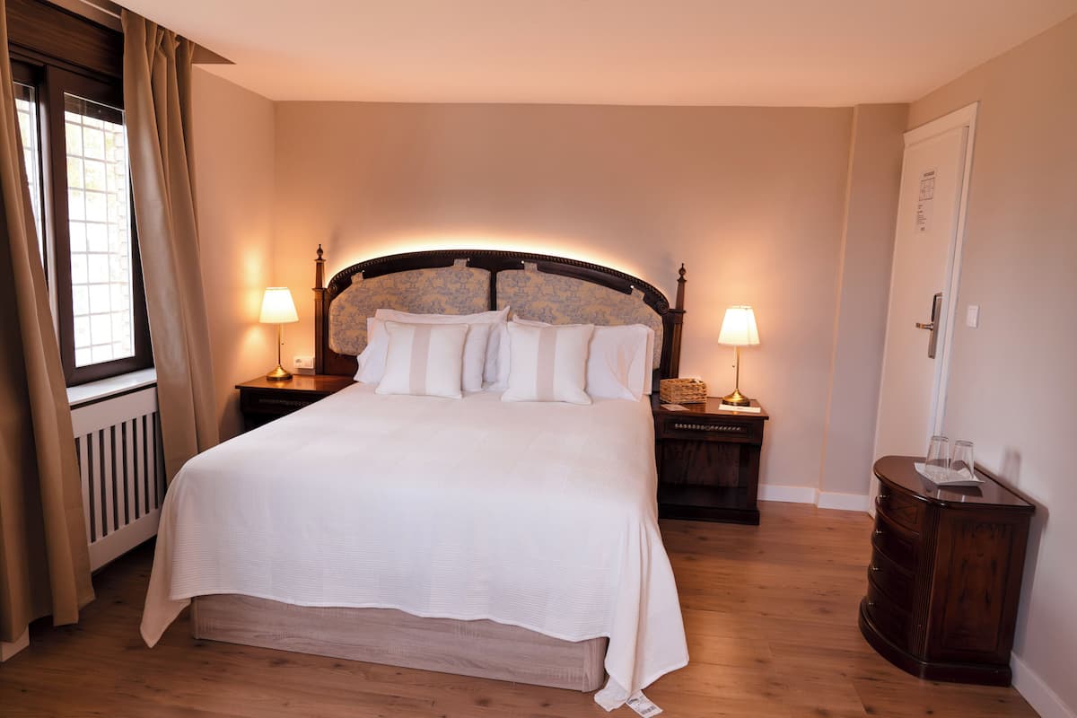 A neatly made bed with a headboard, two bedside tables with lamps, a wooden floor, and a window with curtains.
