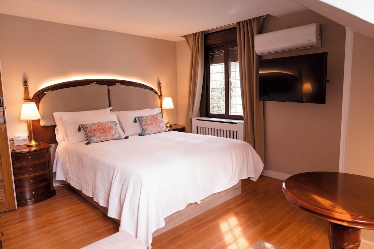 A neatly arranged bedroom with a large bed, bedside lamps, a wooden nightstand, a round wooden table, and an air conditioning unit mounted on the wall.