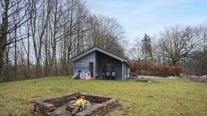 A rustic cabin with a fire pit in the foreground, surrounded by a grassy yard and leafless trees.
