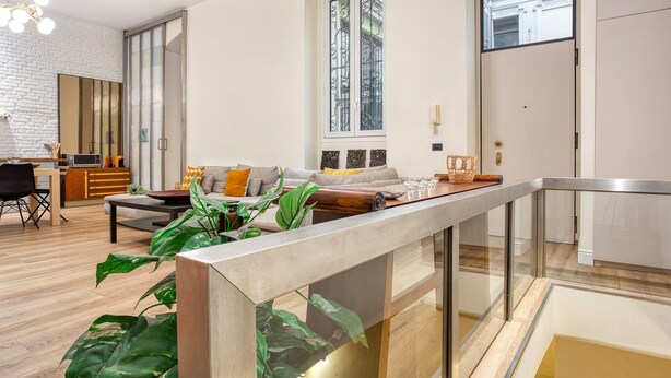 A modern living room with a sofa, coffee table, and large green plants. There is a sliding glass door leading to a balcony, and a dining area with a table and chairs. - JOIVY Stylish Central Maisonette (Milan)