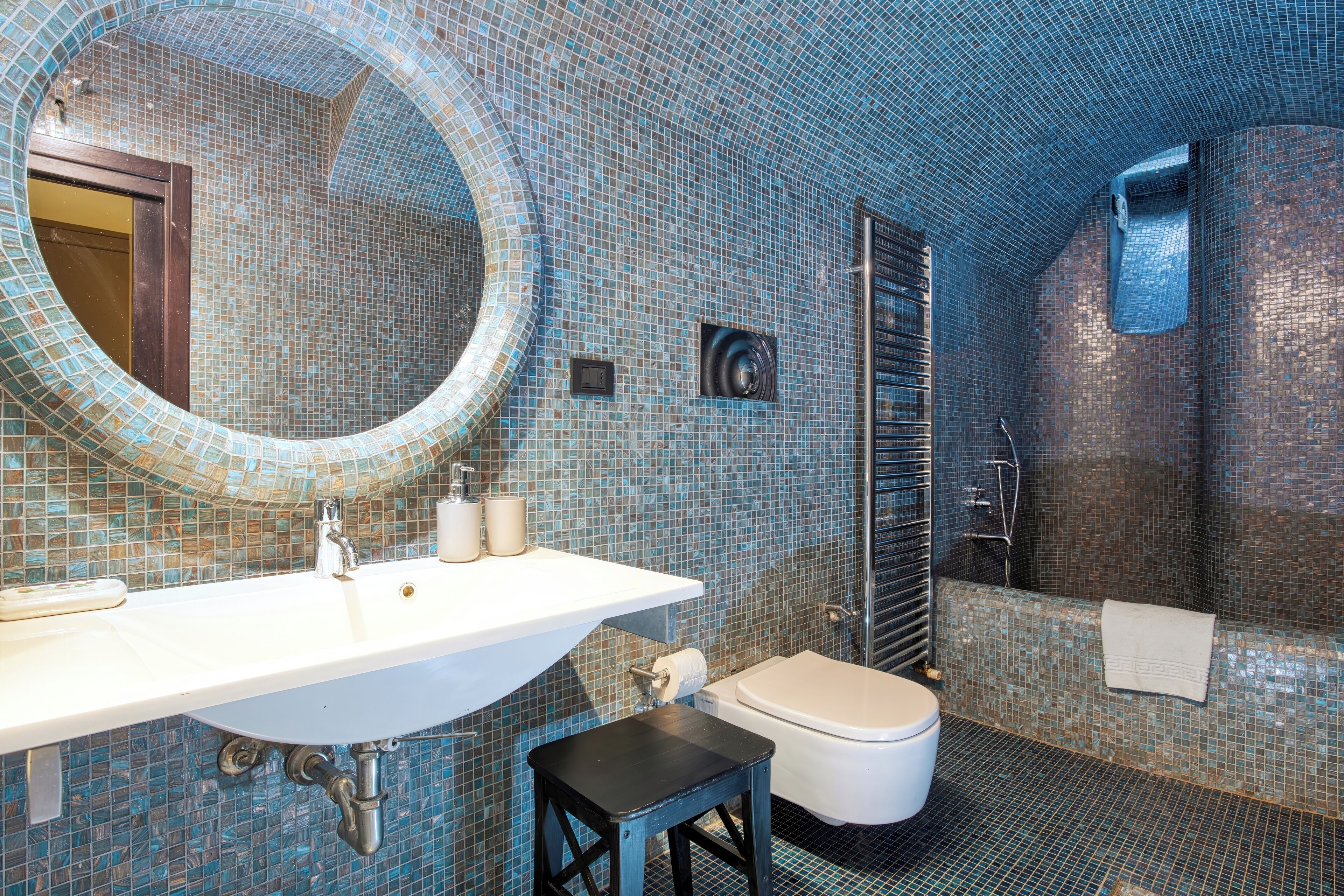 Une salle de bain avec un mur recouvert de mosaïques, un miroir rond, un lavabo, des toilettes et un espace douche.