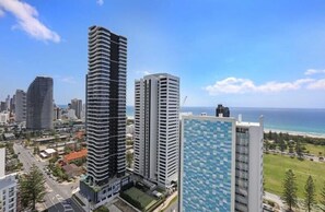 A high-rise building with a curved facade, adjacent to a modern building with a patterned facade, overlooking a coastal area with a clear sky.