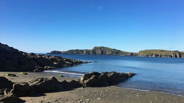 Un rivage rocheux bordé d’une eau calme et dominé par des collines lointaines.