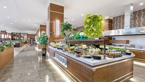 Un moderno vestíbulo de hotel con una recepción, una zona de buffet con diversas opciones de comida y plantas decorativas.