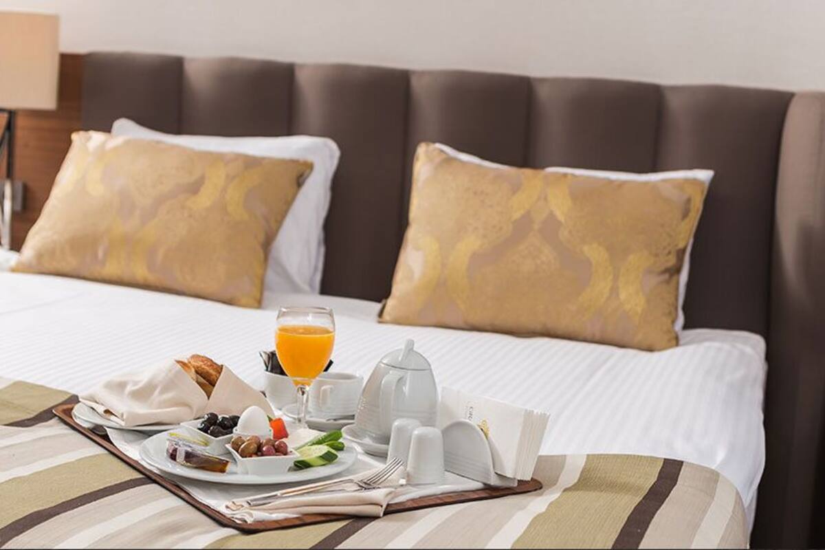 Una cama bien tendida con una bandeja de desayuno, que incluye tostadas, huevos y jugo de naranja, en una habitación de hotel con cabecera marrón y almohadas estampadas.