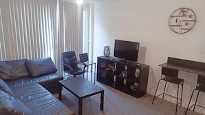 Un salon avec un canapé en cuir, une table basse, un téléviseur posé sur un meuble et un petit bureau avec deux chaises.