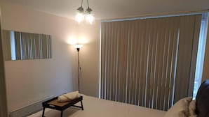Une chambre d’hôtel comprenant un lit, une table de chevet, une lampe et une grande fenêtre avec des stores.