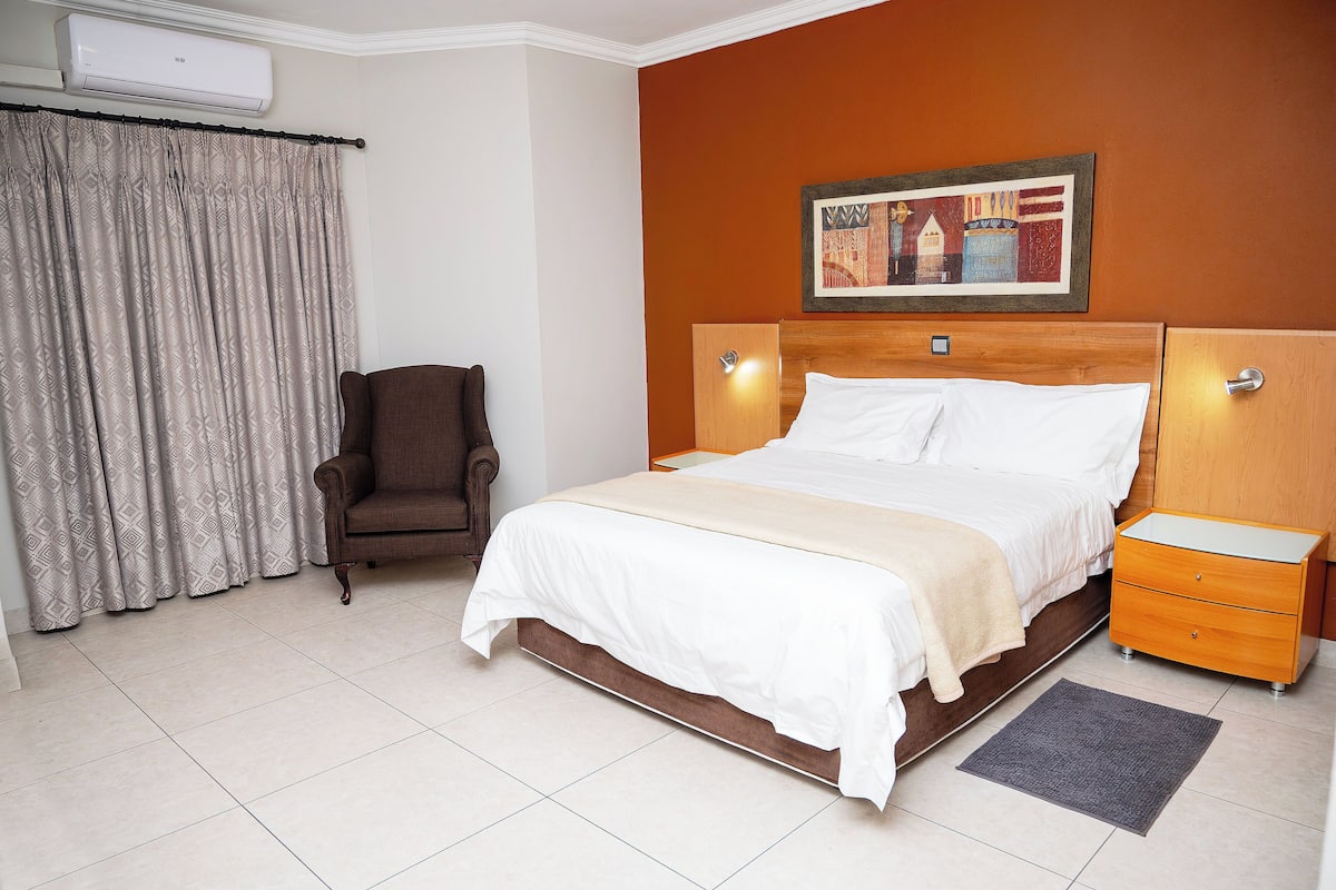 A hotel room with a bed, a chair, a nightstand, and a painting on the wall.