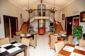 A spacious dining area with wicker furniture and a central round table, a staircase with a railing, and a reception area with a sign. - Boksburg Boutique Hotel (Boksburg)