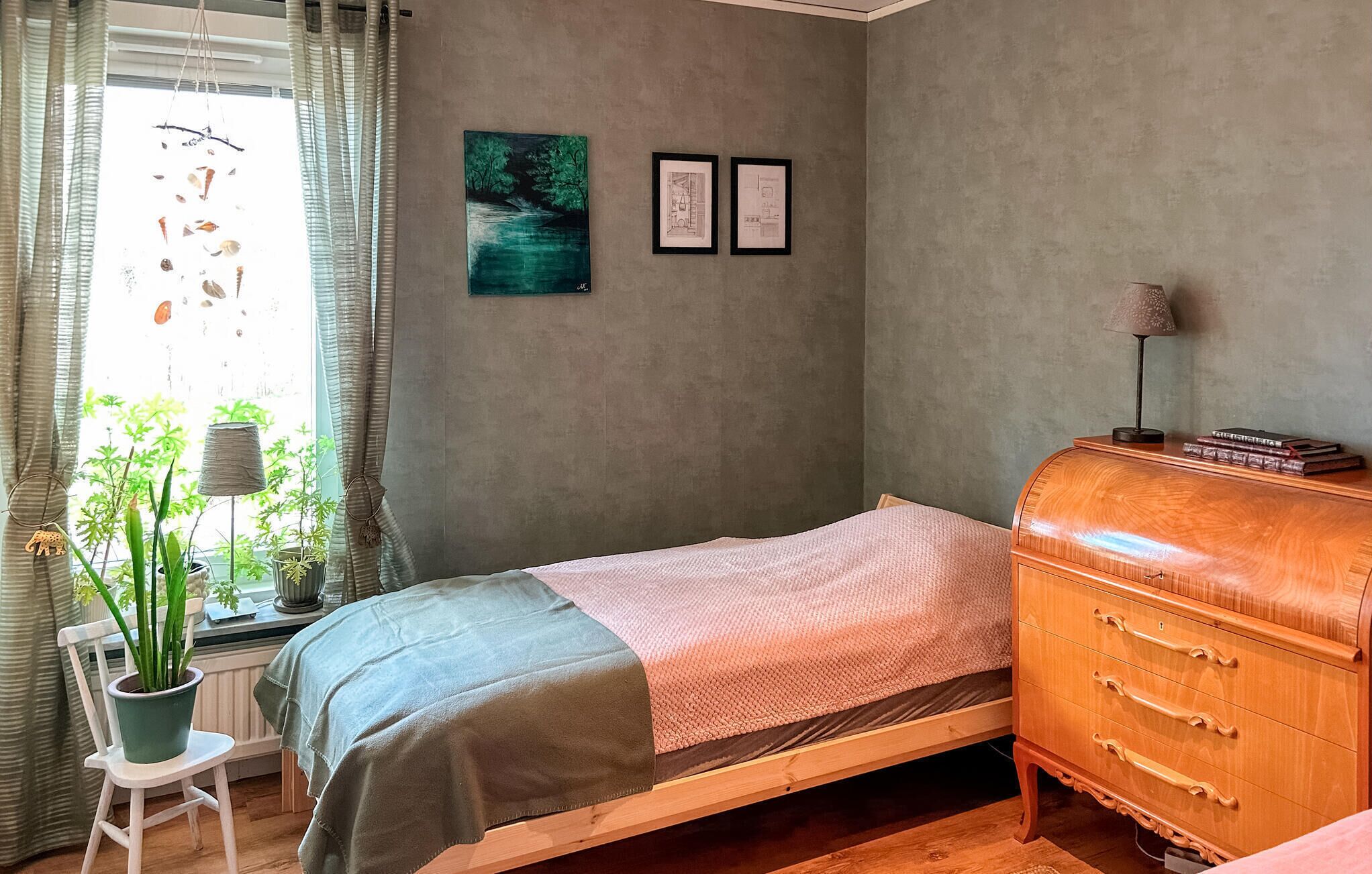 Une chambre à coucher avec un lit, une commode, une fenêtre avec des rideaux et deux tableaux encadrés accrochés au mur.