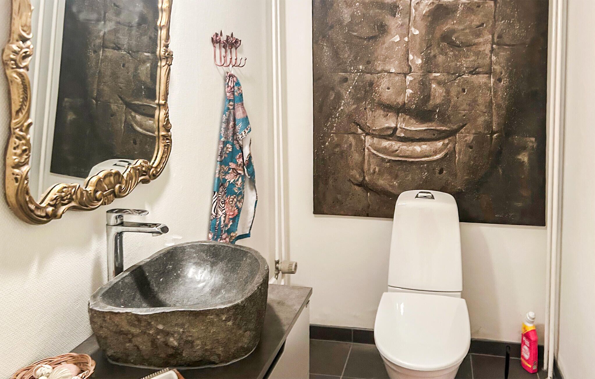Een badkamer met een stenen wastafel, een wit toilet, een spiegel met een sierlijk frame en twee aan de muur bevestigde kunstwerken.
