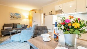 A modern living room with a sofa, a dining table with a vase of flowers, and a kitchen area in the background.