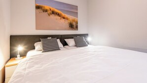 A neatly made bed with white linens and two pillows, a nightstand with a lamp, and a painting of a beach scene on the wall.