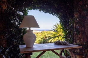 Una mesa con una lámpara blanca y un florero, frente a un fondo de follaje y vista al mar.