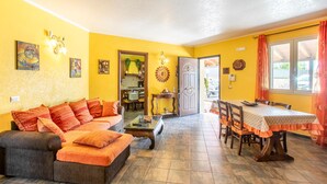 A living room with a sofa, coffee table, and dining table. There is a kitchen visible through an open door.