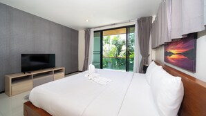 A modern hotel room with a large bed, a flat-screen TV on a wooden stand, and a balcony with a view of greenery. - Pool villa in rawai  (Tambon Rawai)